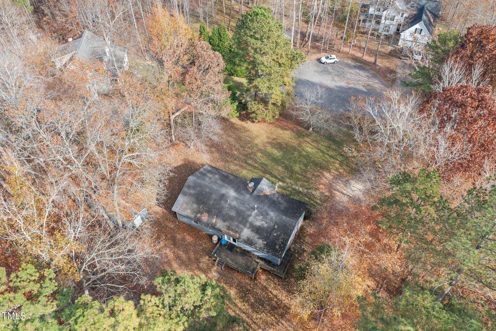 Aerial view of a solitary house with a black roof, surrounded by trees with autumn-colored leaves, situated near a road with adjacent houses visible in a suburban area.