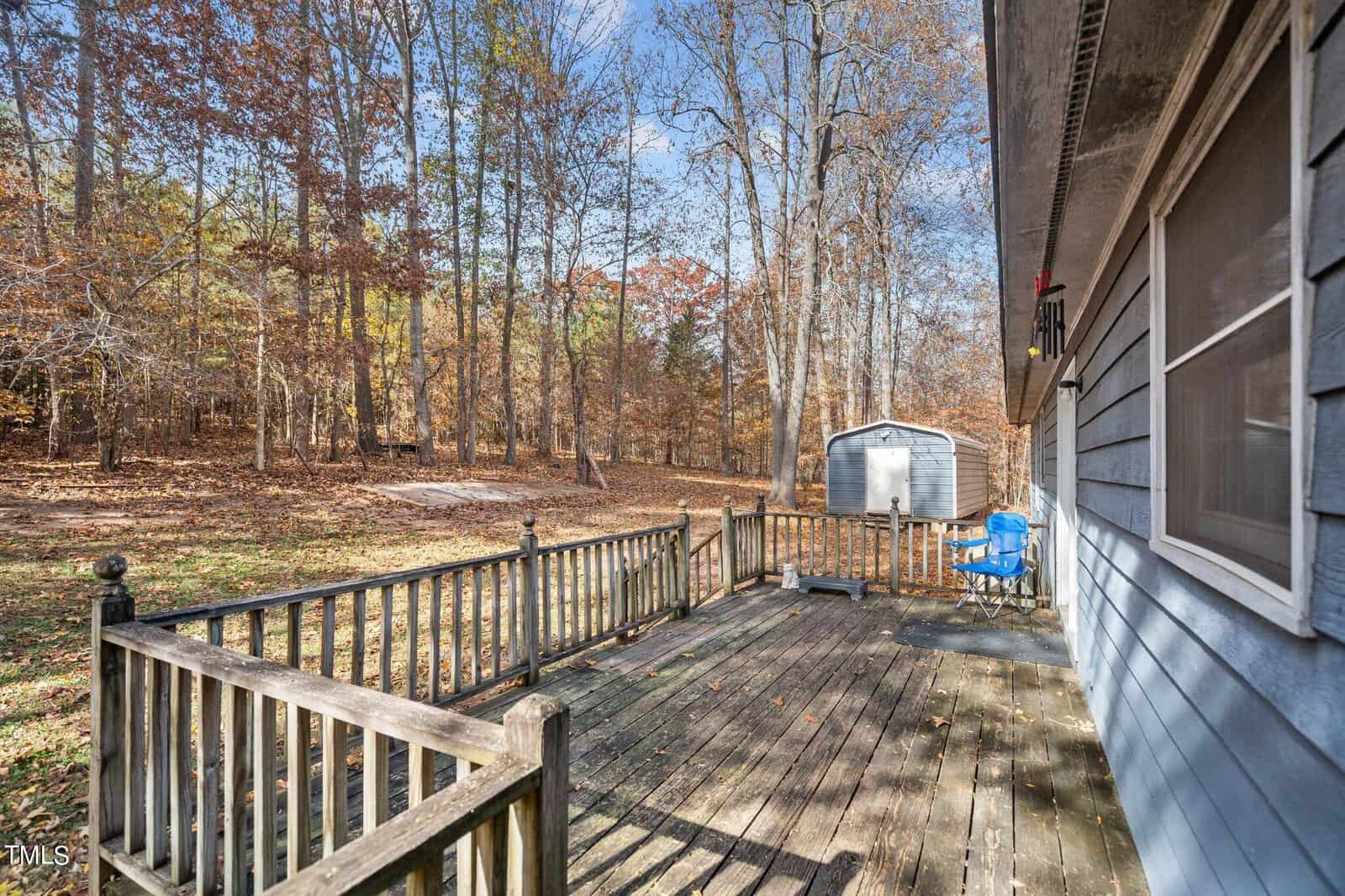 A wooden deck attached to a house, overlooking a backyard with leafless trees and a shed. the deck features a weathered railing, and there is a blue child's chair visible. autumn leaves cover the ground.