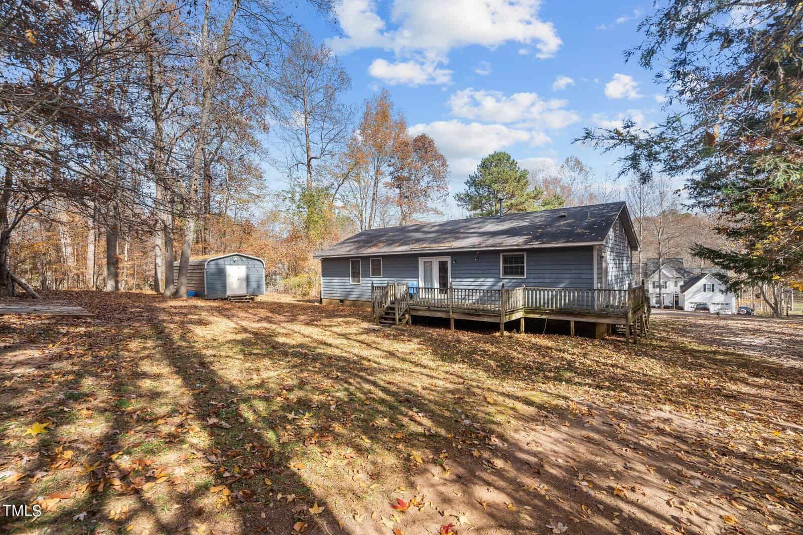 A single-story wooden house on stilts with a metal shed and bare trees around. sunlight casts long shadows across the leaf-covered ground. no visible people or movement.