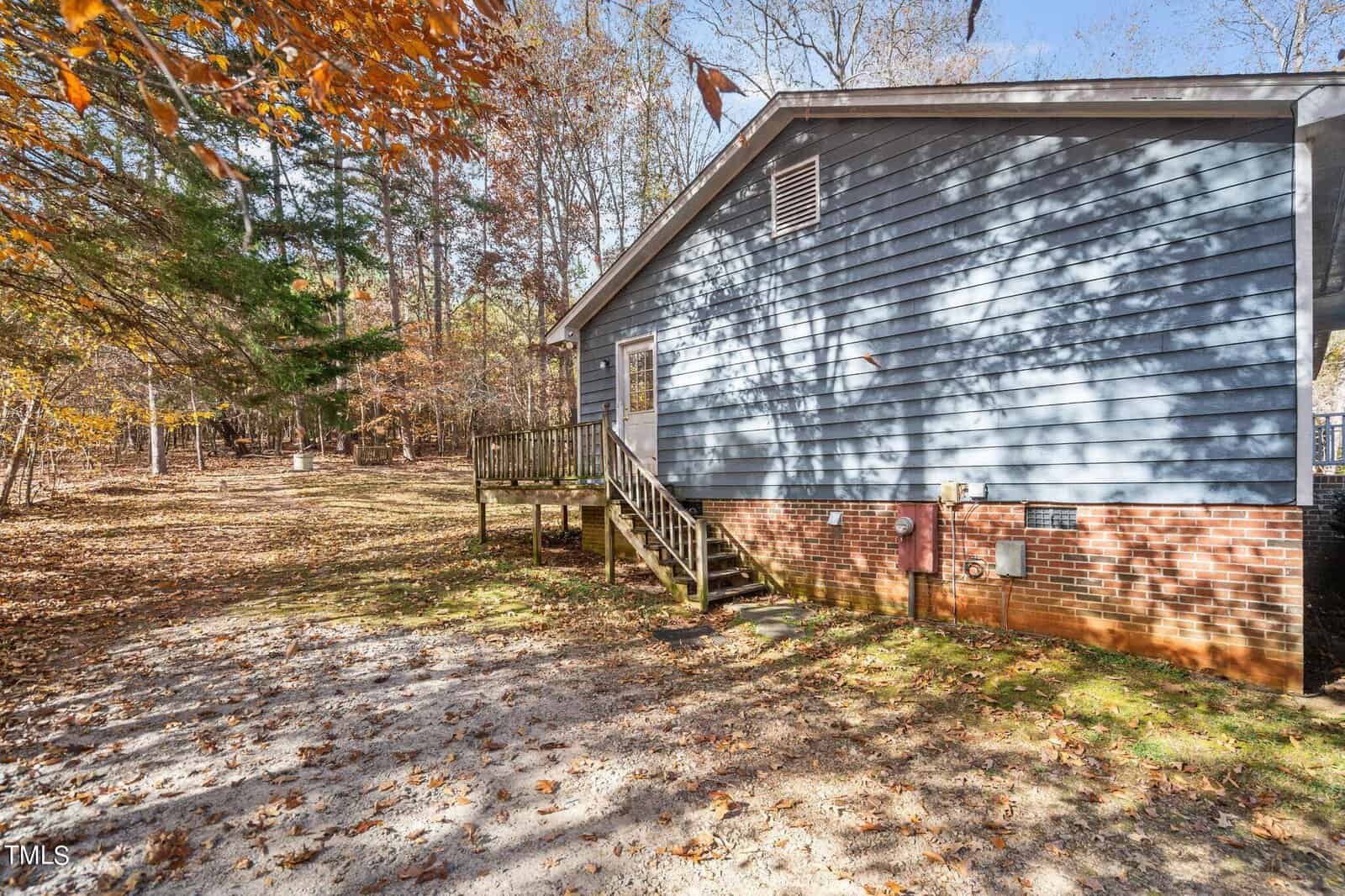 Side view of a blue-sided house with a wooden deck surrounded by a leaf-covered yard and trees in autumn. bright sunlight casts shadows on the house.