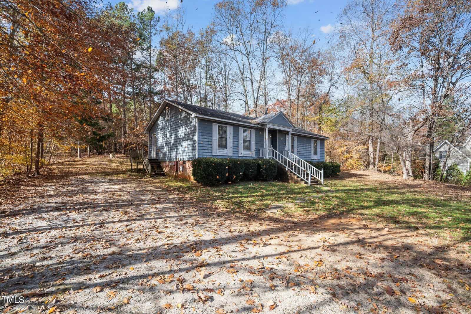 A single-story home with gray siding and white trim, featuring a front deck and surrounded by a leafy yard with scattered autumn leaves, under a partly cloudy sky.