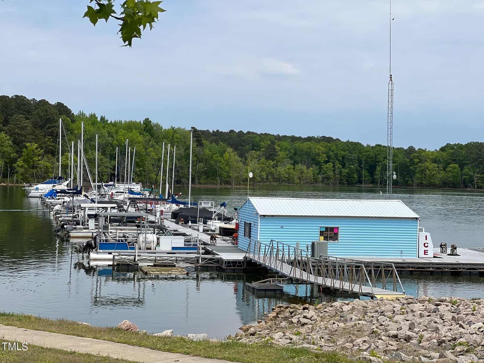 A scenic view of a marina with various sailboats docked alongside a floating blue building, surrounded by a tranquil lake and a lush treeline under a cloudy sky.