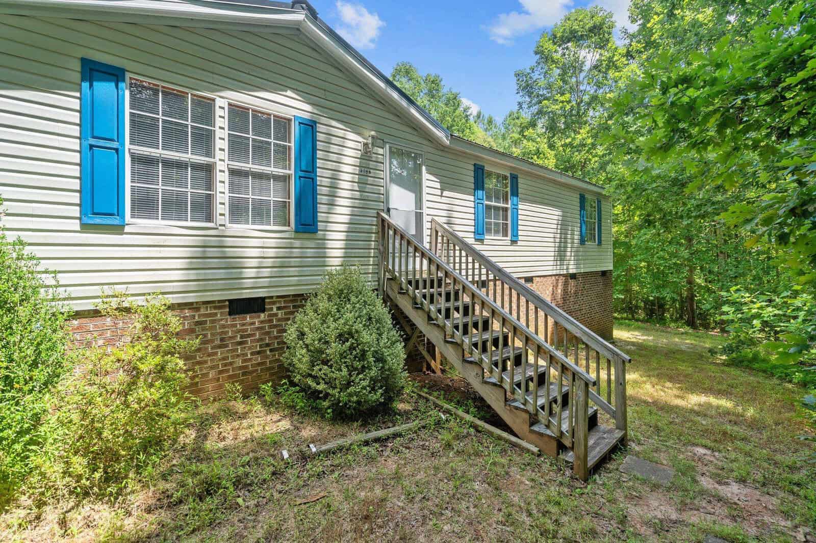 A single-story house with beige siding and blue shutters, featuring a front porch with stairs leading to the entrance. The surrounding greenery includes trees and shrubs, and the sky is clear with a few clouds.