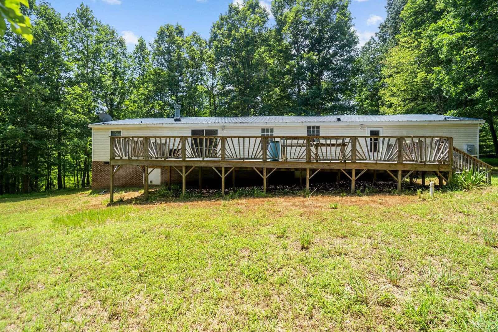 A long mobile home with a wraparound wooden deck is set on a grassy lawn. Tall trees surround the property, and the sky is clear and blue.