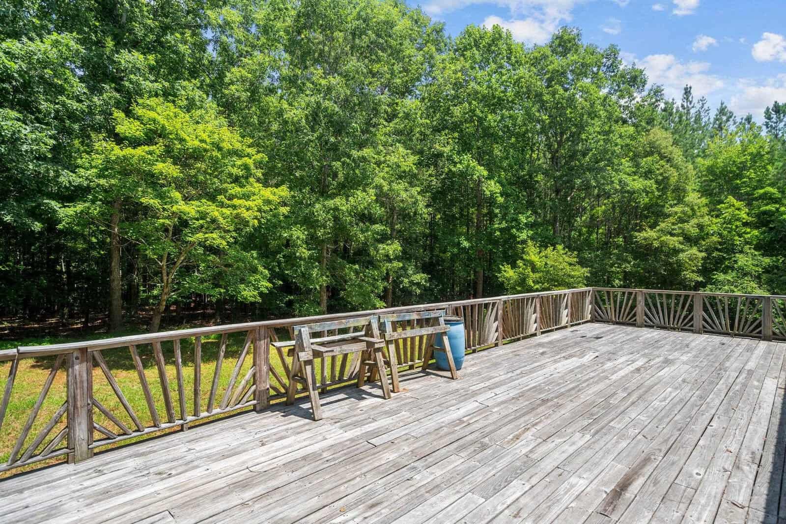A spacious wooden deck overlooking a lush, green forest under a blue sky. A few chairs and a barrel are arranged along the railings, providing a serene outdoor setting. The trees in the background create a tranquil natural ambiance.