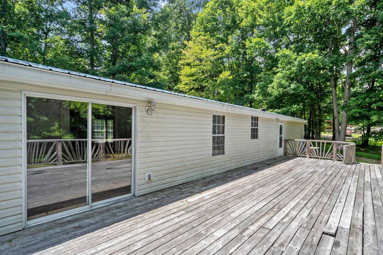 A long, single-story white mobile home with several windows and a sliding glass door opens onto a large wooden deck. The deck is surrounded by lush green trees.