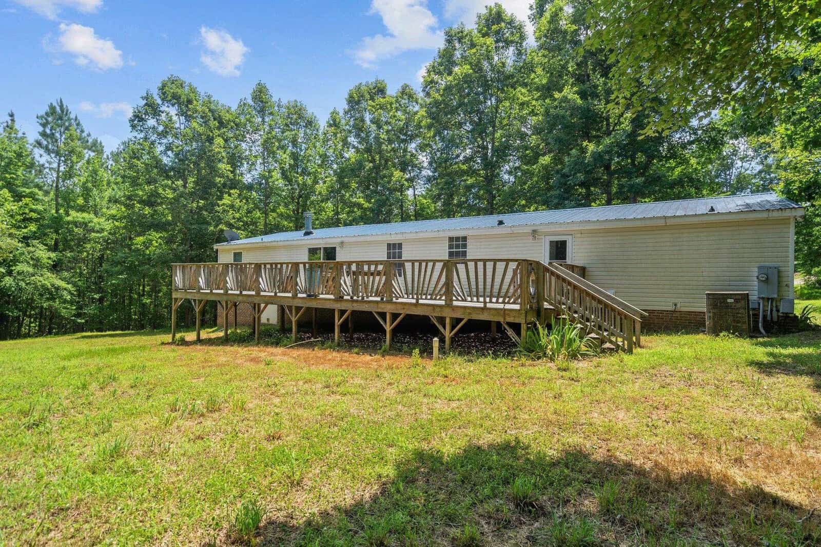 A white mobile home with a metal roof and a large wooden deck on a grassy yard surrounded by tall trees under a partly cloudy sky.