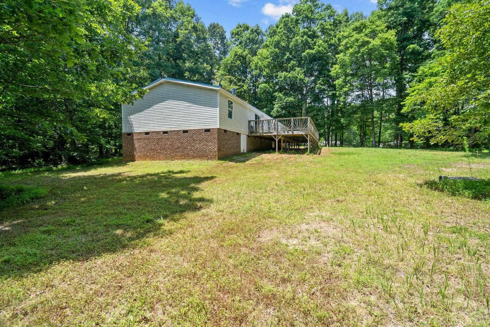 A house with a raised foundation and a side deck is situated in a grassy yard. The property is bordered by lush green trees under a blue sky with scattered clouds.