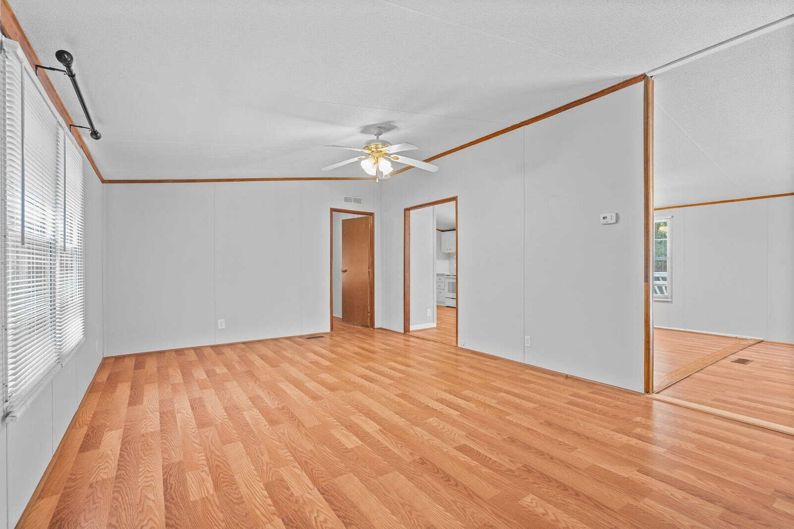An empty room with wood flooring and light gray walls. A ceiling fan with lights hangs from the ceiling. There are two open doorways and large windows with blinds, allowing natural light to illuminate the space.