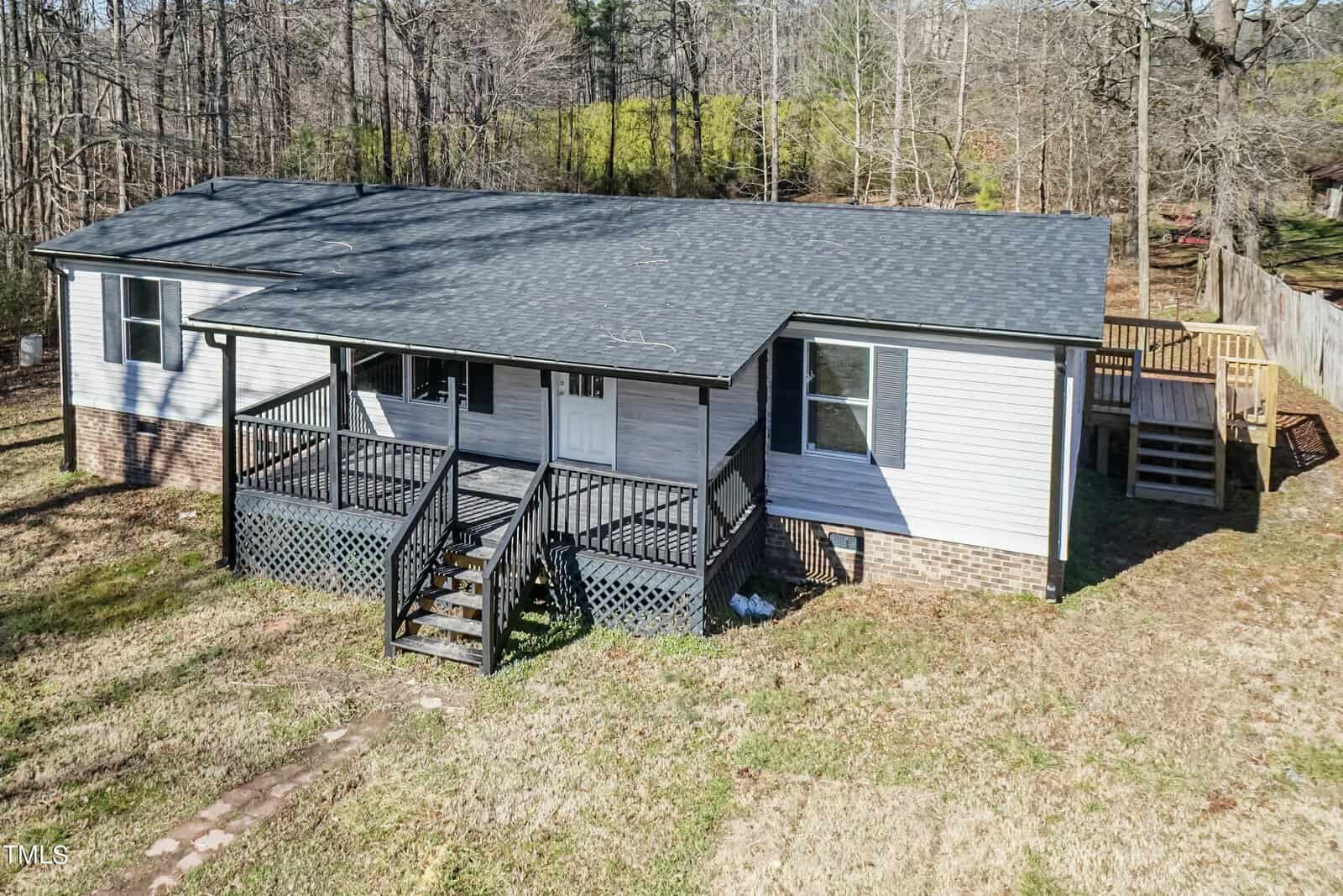 Aerial view of a single-story house with white siding, a gray shingled roof, and three sets of stairs leading to separate entrances. the home is surrounded by a leafless wooded area.