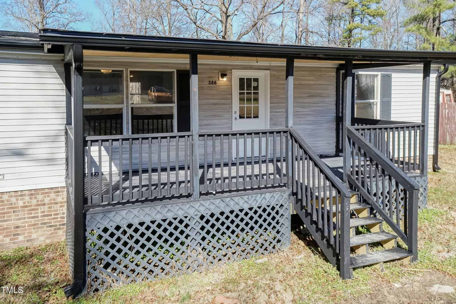 A single-story house with a covered front porch featuring a wooden deck and stairs, surrounded by a railing and lattice skirting. the house has white siding and black shutters.