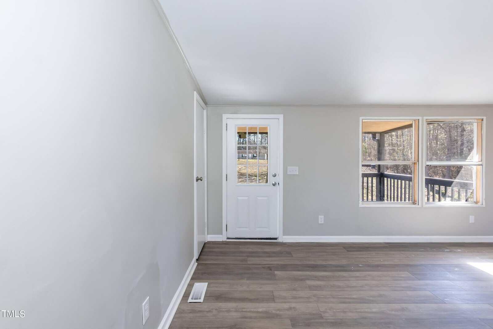 Empty living room interior with gray walls, wood flooring, a closed white door, and multiple windows letting in natural light. the room is unfurnished and freshly painted.