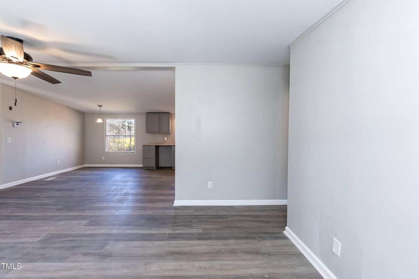 An empty, brightly lit room featuring gray laminate flooring, white walls, and a ceiling fan. in the background, there is a glimpse of a kitchen area.