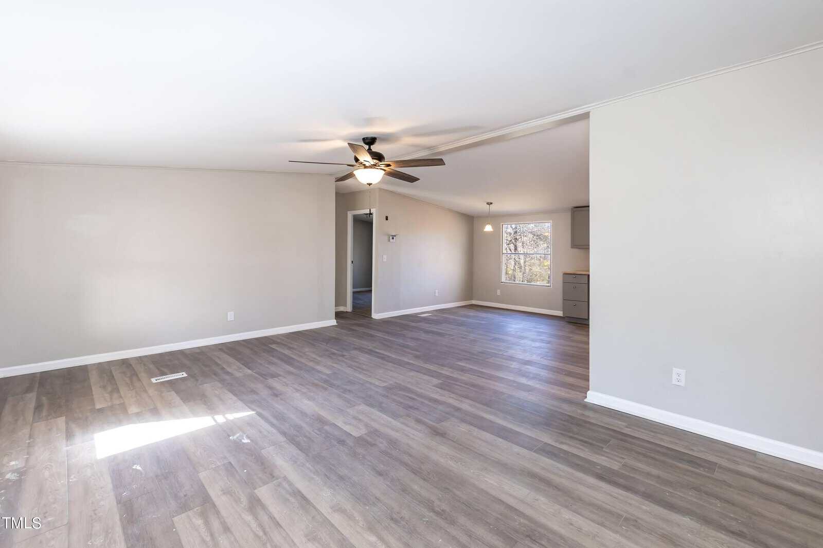 An empty living room featuring grey wood flooring, white walls, and a ceiling fan. a window provides natural light. the room transitions to another area with a kitchen visible in the background.