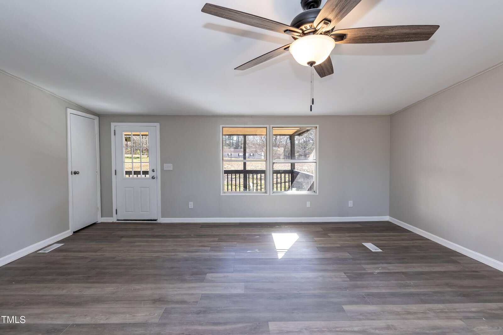 Empty living room with a wooden floor, white walls, and a ceiling fan. rear wall features a door and large window overlooking a deck or patio.