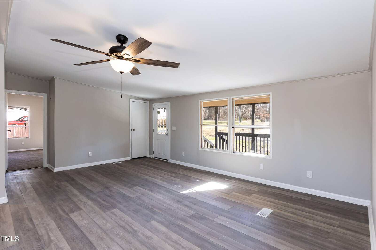Spacious and empty room with laminate flooring, featuring large windows, a ceiling fan, and an open back door leading to an outdoor deck. walls are light-colored, and the space is brightly lit by natural light.