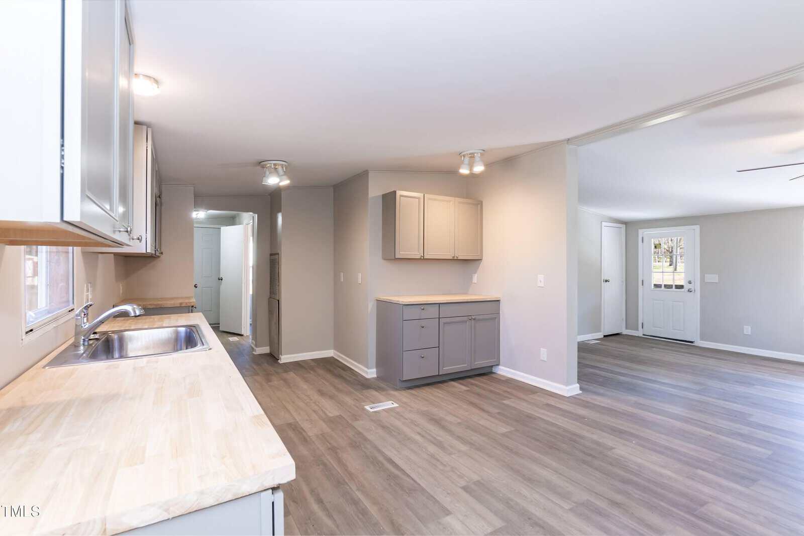 Empty kitchen and living area in a newly renovated home, featuring modern appliances and light wood floors, with grey and beige cabinets and white walls.