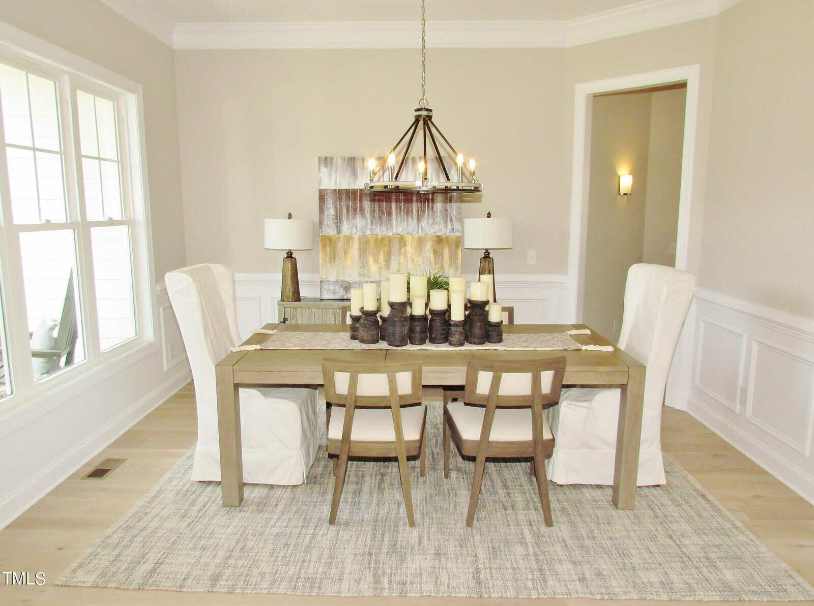 A modern dining room with a rectangular wooden table, six chairs, two white slipcovered armchairs at each end, a chandelier, decorative candles, two table lamps, and abstract wall art. The room has light walls and floors.