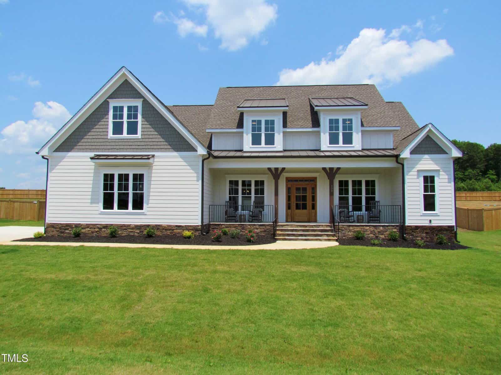 A modern two-story house with gray siding, gabled roofs, large windows, and a covered front porch with chairs, set on a spacious green lawn under a blue sky with a few clouds.