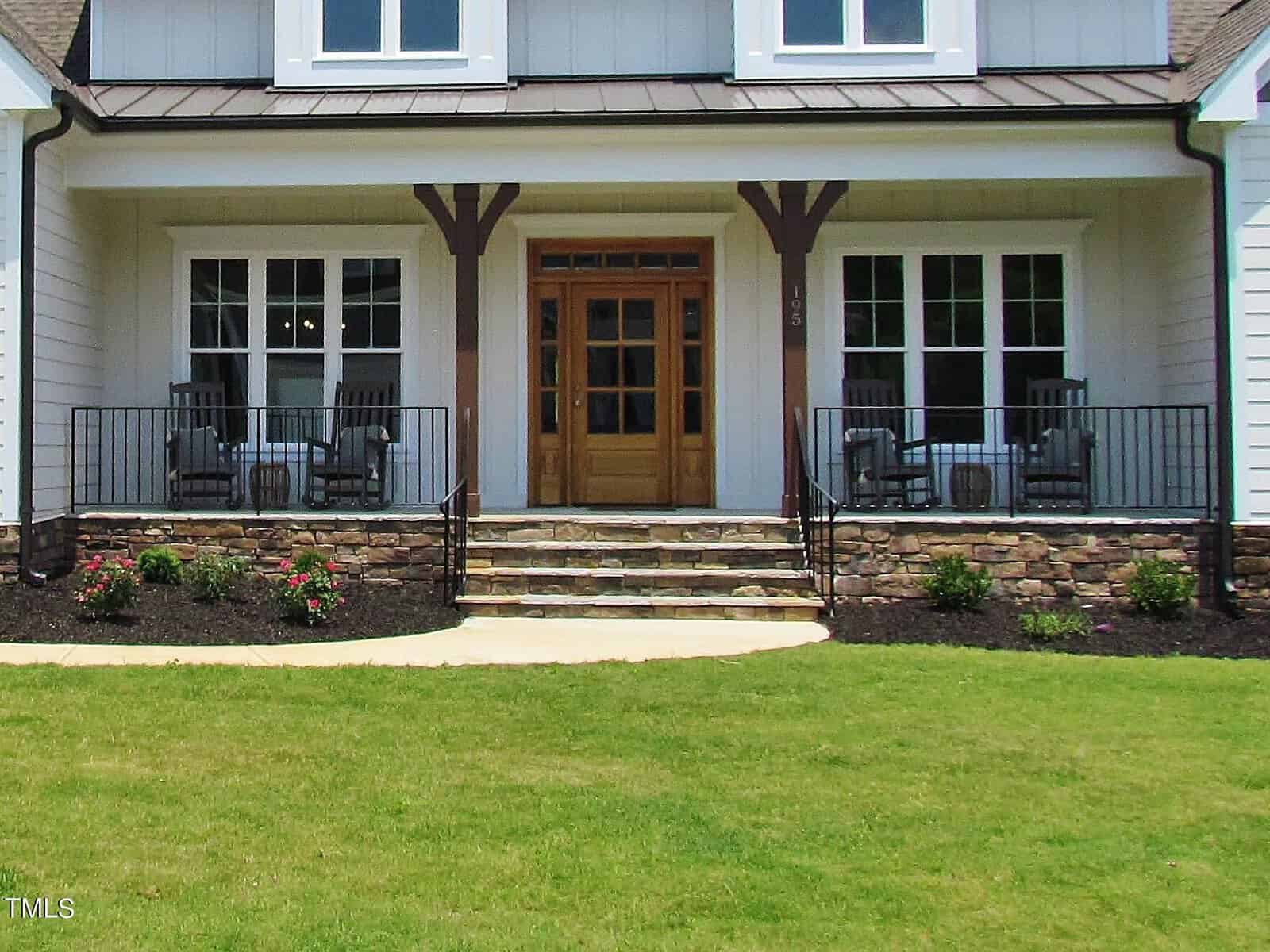 A house with a wooden double front door, two large windows on each side, a stone porch with steps, black railings, rocking chairs, and a well-kept lawn with flower beds.