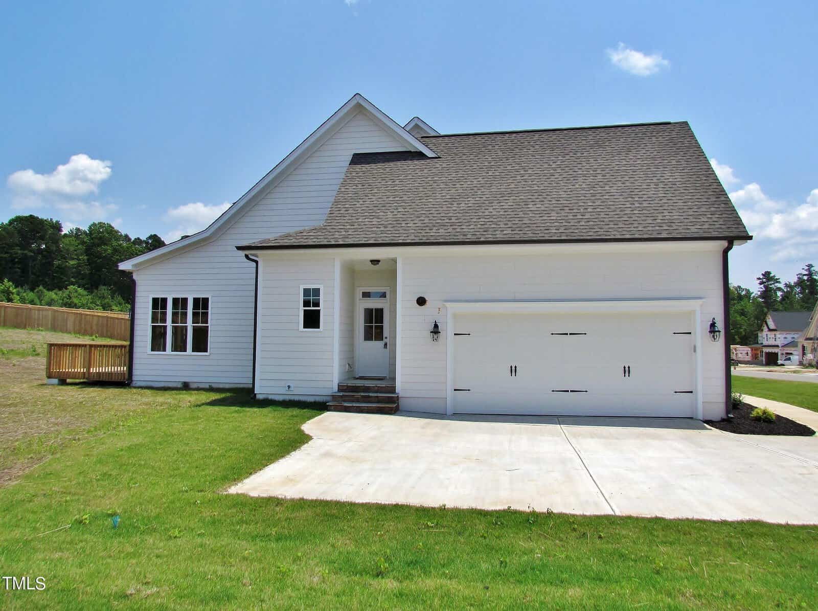 A modern white house with black roof shingles, a two-car garage, and a grassy lawn sits on a sunny day with a blue sky and light clouds. There is a wooden deck on the left side and a driveway leading to the garage.