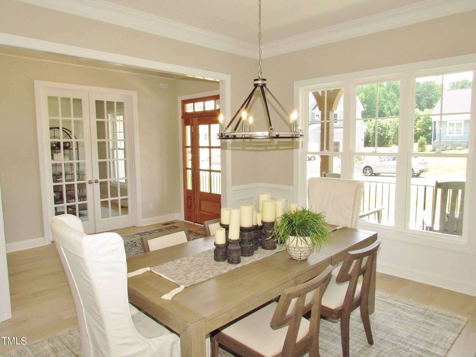 A bright dining room with a wooden table, six chairs, a modern chandelier, and a centerpiece of candles and greenery. Large windows let in natural light, and a front door and French doors are visible in the background.
