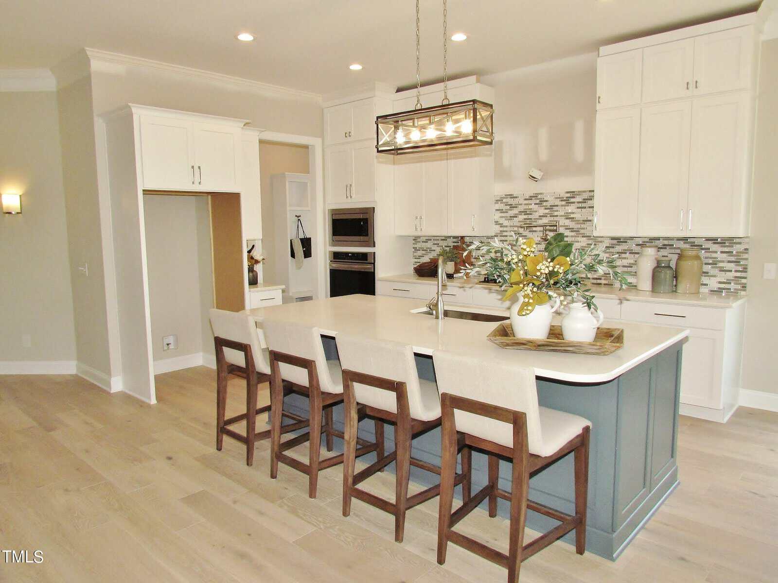 Modern kitchen with light wood floors, a large island with seating for five, white cabinets, stainless steel appliances, mosaic tile backsplash, and a decorative vase with flowers on the island.