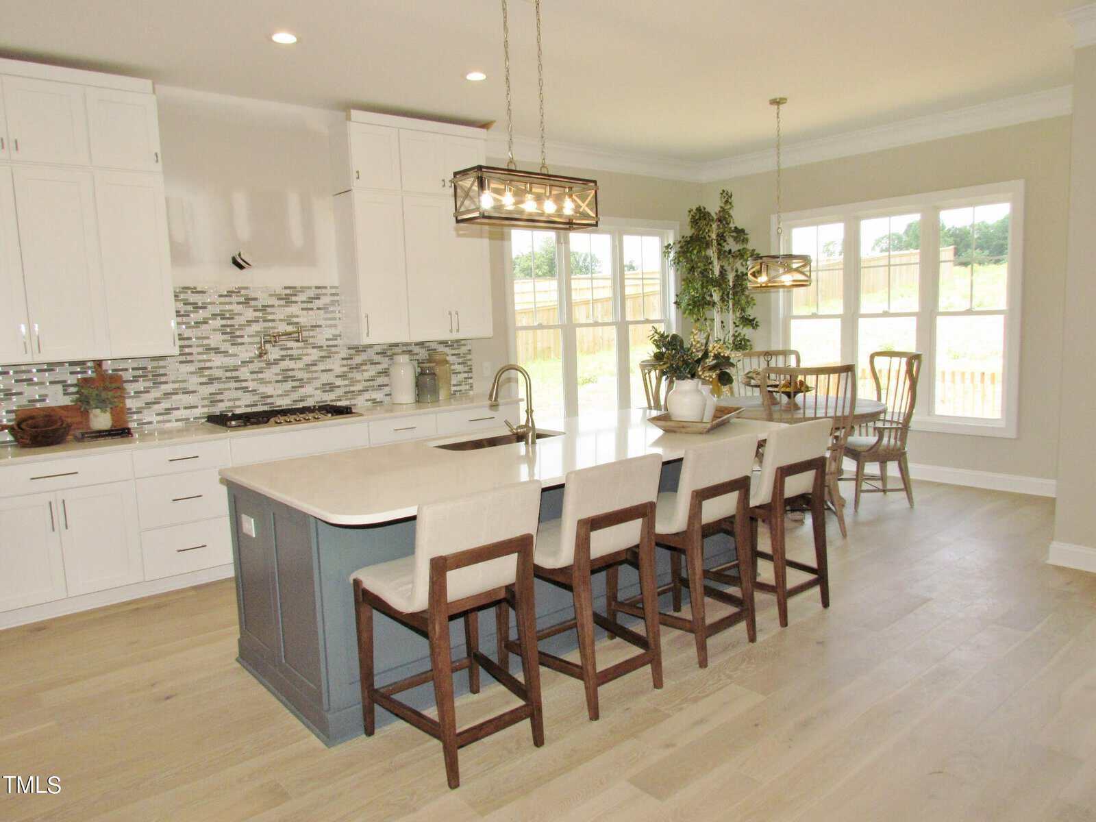 Modern kitchen with white cabinets, a large island with sink and four chairs, mosaic tile backsplash, pendant lights, and a dining area with a table and chairs near large windows.