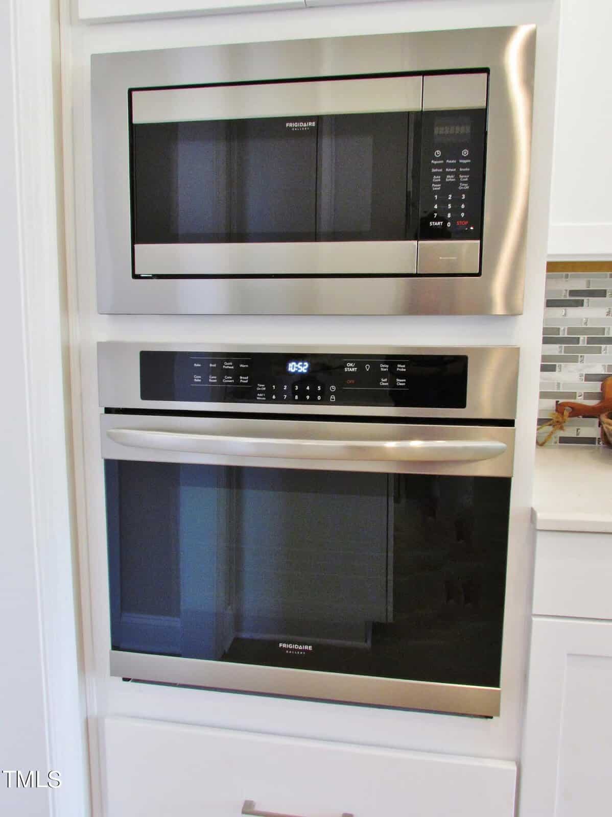 Built-in stainless steel microwave above a built-in wall oven, both with digital displays and touch controls, set into white cabinetry next to a kitchen counter with a glass tile backsplash.