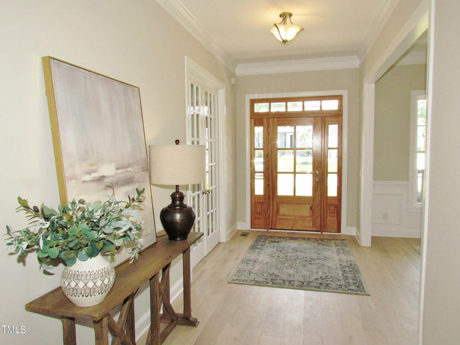 A bright entryway with light wood floors, a patterned rug, a glass-paneled wooden front door, and a console table decorated with a lamp, vase, and greenery. Framed art hangs above the table.