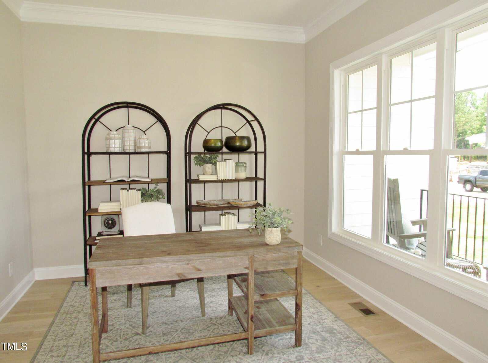 A bright home office with a wooden desk and chair, a small plant on the desk, two black metal bookshelves with books and decor, beige walls, and large windows letting in natural light.