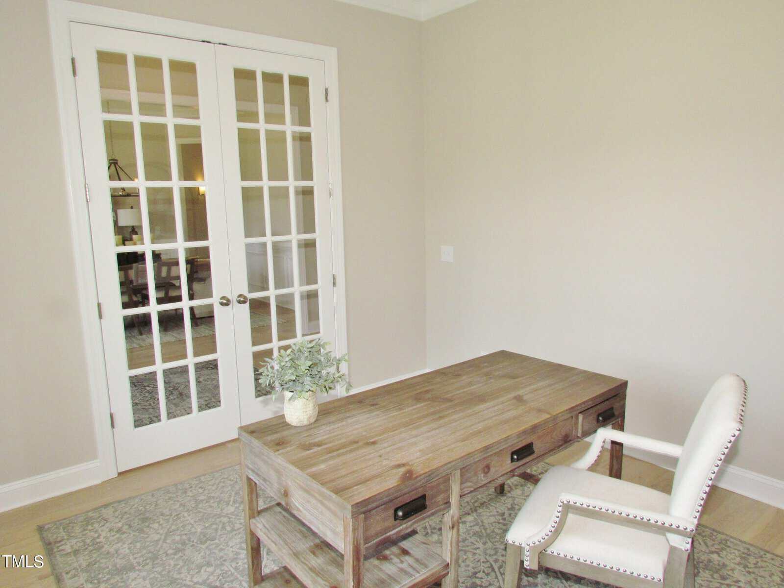 A simple home office with a wooden desk, a white upholstered chair, a small potted plant on the desk, light walls, and double glass French doors leading to another room.