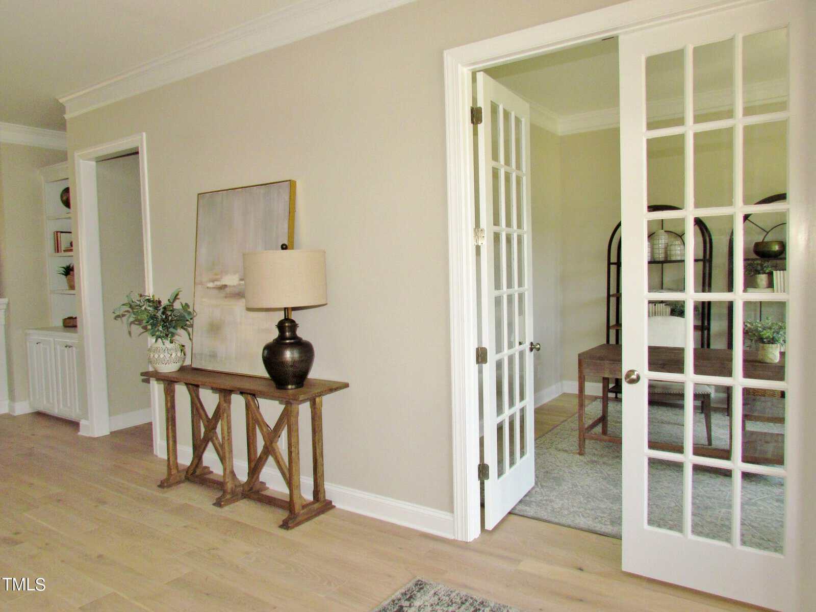 A bright room with light wood floors, a rustic console table with a lamp and plant, and an open French door leading to a home office with a desk and bookshelf. Walls are painted a soft beige.