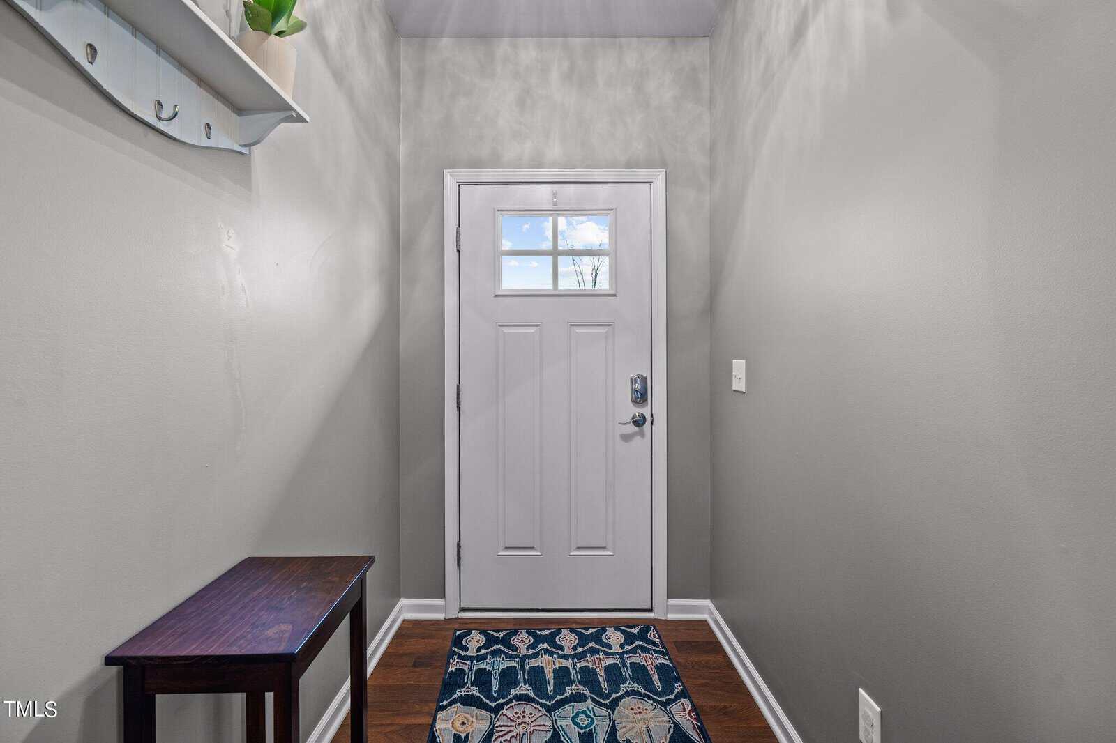 A narrow, light-gray entryway with a white door featuring a small window. The floor is dark wood with a colorful patterned rug. A dark wooden bench sits on the left, and a white shelf with a potted plant hangs above.