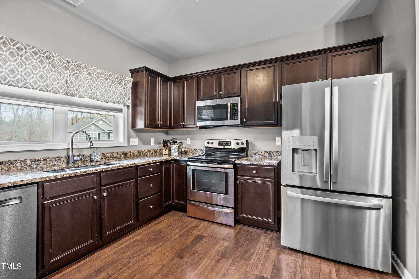 Modern kitchen featuring dark wood cabinets, stainless steel appliances, and granite countertops. A large window above the sink provides natural light, with a patterned valance. The room has hardwood flooring.