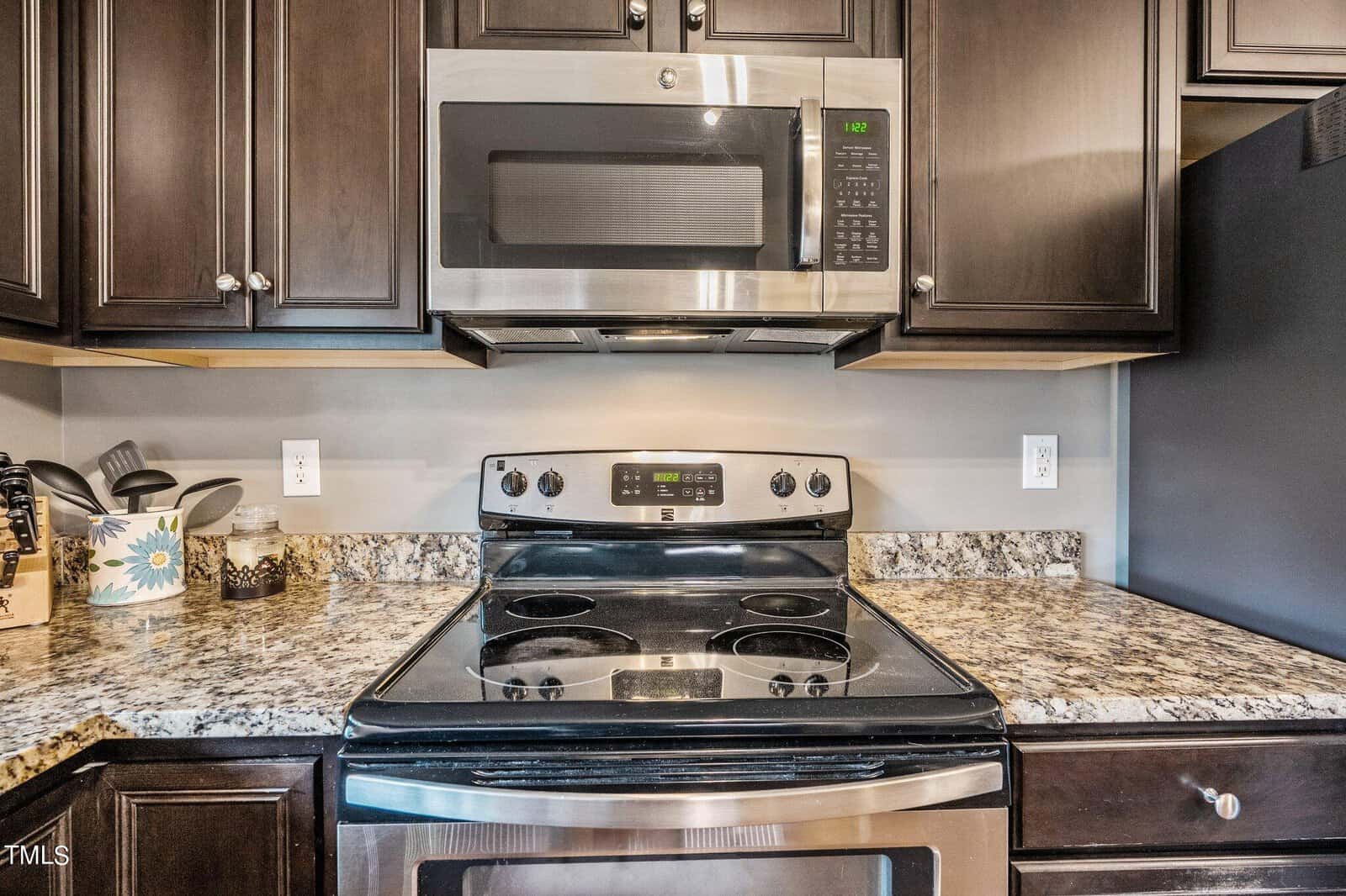 A modern kitchen with dark wood cabinets, a stainless steel oven, and a mounted microwave. The countertop is speckled granite, with kitchen utensils and a plant. The clock on the microwave reads 1:02.