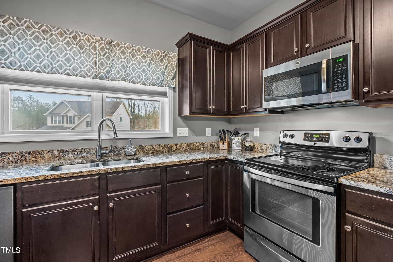 Modern kitchen with dark wood cabinets, granite countertops, and stainless steel appliances, including an oven and microwave. A window above the sink has patterned curtains and provides a view of a house and trees outside.