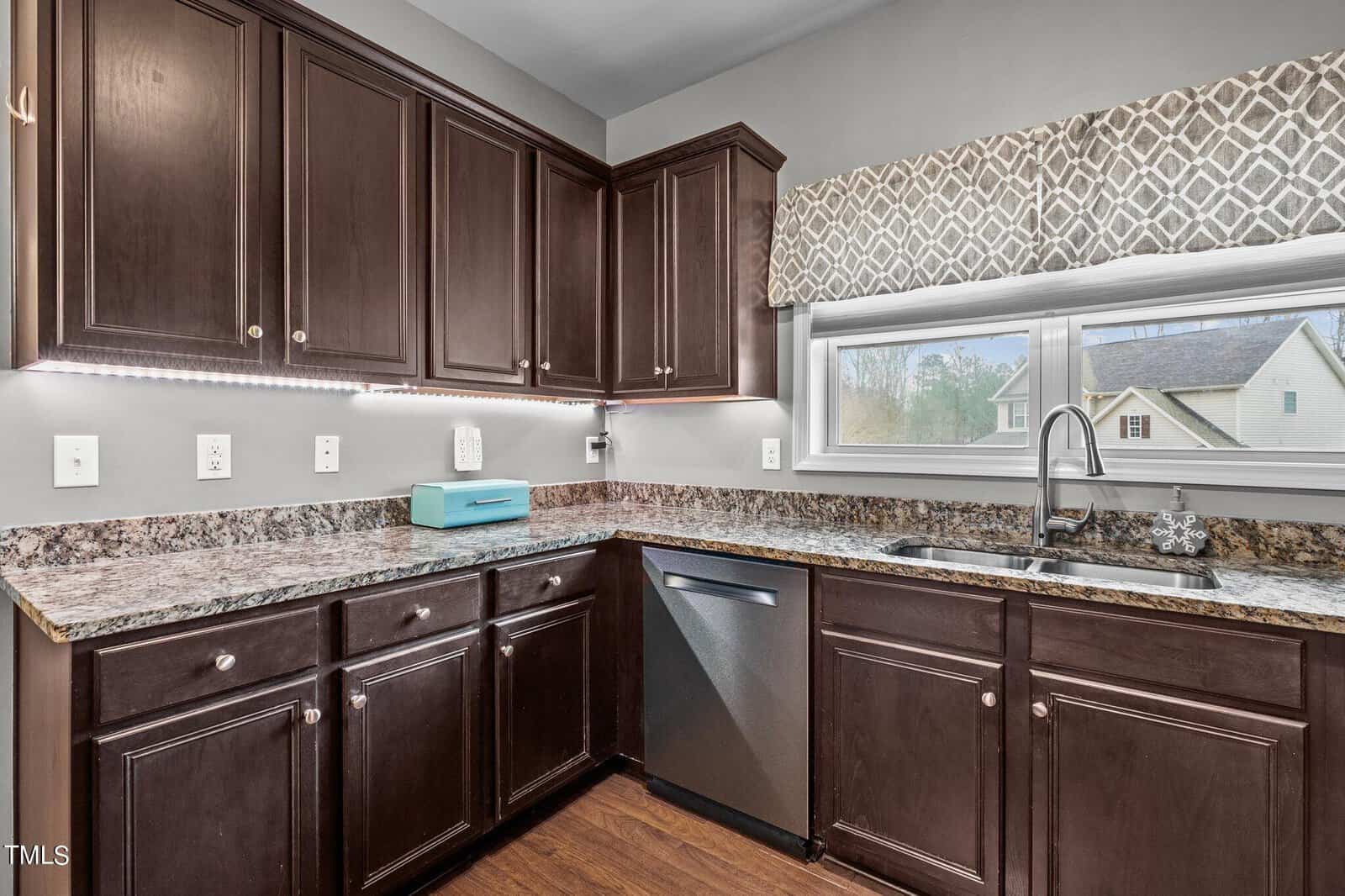 A modern kitchen with dark wood cabinets, granite countertops, and stainless steel appliances, including a dishwasher. Under-cabinet lighting illuminates the counter. A window above the sink has a patterned valance, and there is a blue microwave on the counter.