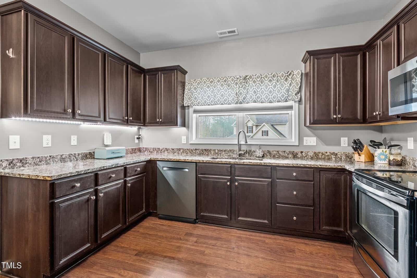 A modern kitchen with dark wooden cabinets, granite countertops, and stainless steel appliances. The space features a large window with a patterned valance, hardwood floors, and under-cabinet lighting.