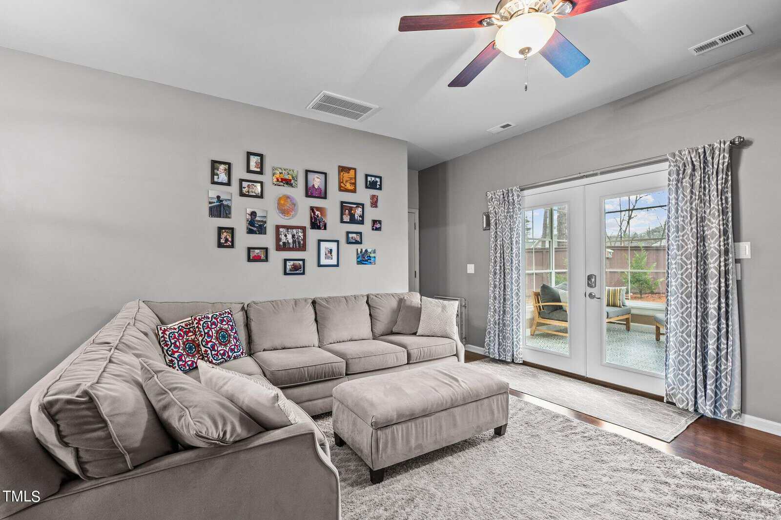 A cozy living room features a large gray sectional sofa and matching ottoman on a white rug. The wall displays several framed photos. Glass doors lead to a patio with outdoor furniture. A ceiling fan with a light is overhead.