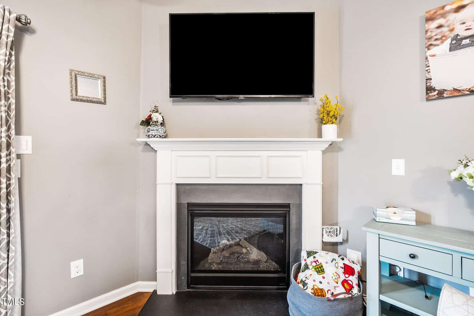A cozy living room corner with a modern fireplace and a mounted flat-screen TV above it. A small decorative shelf on the mantel holds flowers and a snowman figurine. A basket with colorful items sits beside a light blue cabinet on the right.