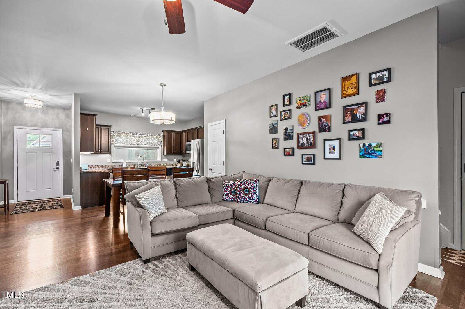 A cozy living room with a beige sectional sofa and a cushioned ottoman. A gallery wall displays numerous family photos. The open-concept space leads to a kitchen with dark wood cabinets and a white dining table. A ceiling fan is above the sofa.