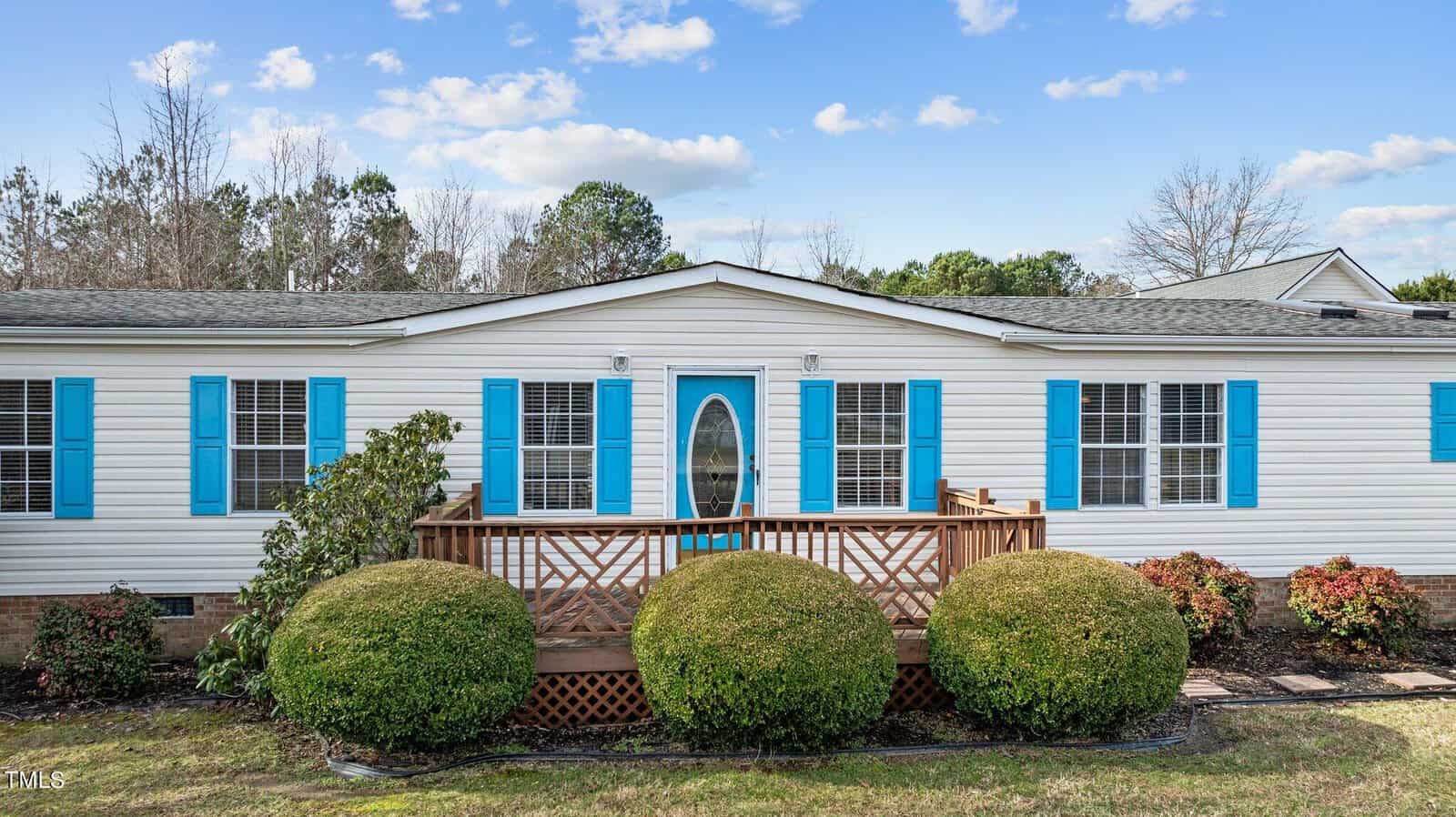 A white single-story house with blue shutters, an oval glass front door, a small wooden porch, and neatly trimmed bushes in the front yard under a blue sky with scattered clouds.