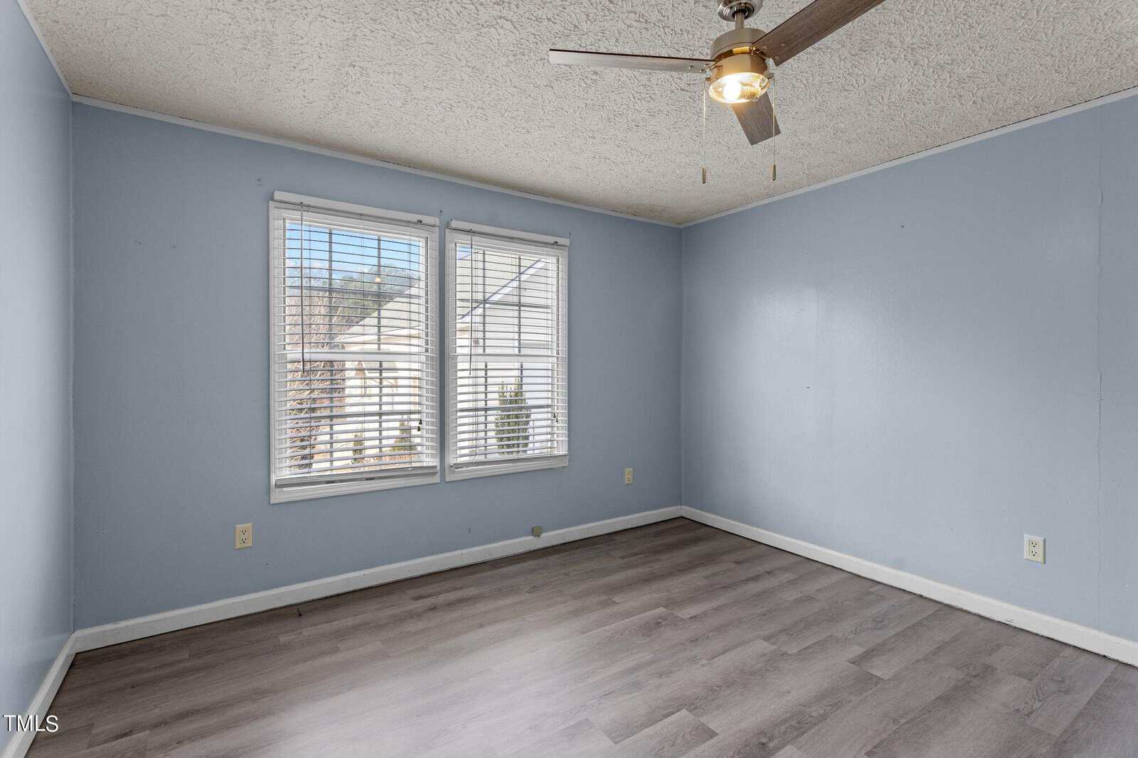 Empty room with light blue walls, two windows with blinds, a ceiling fan with a light, and gray wood-style flooring. Sunlight comes through the windows, revealing part of a neighboring house outside.