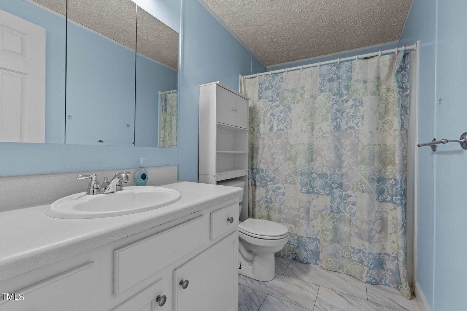 A bathroom with light blue walls, a white vanity with a sink and mirror, a white toilet, shelving unit, and a bathtub with a floral-patterned shower curtain. The floor has white and gray tiles.