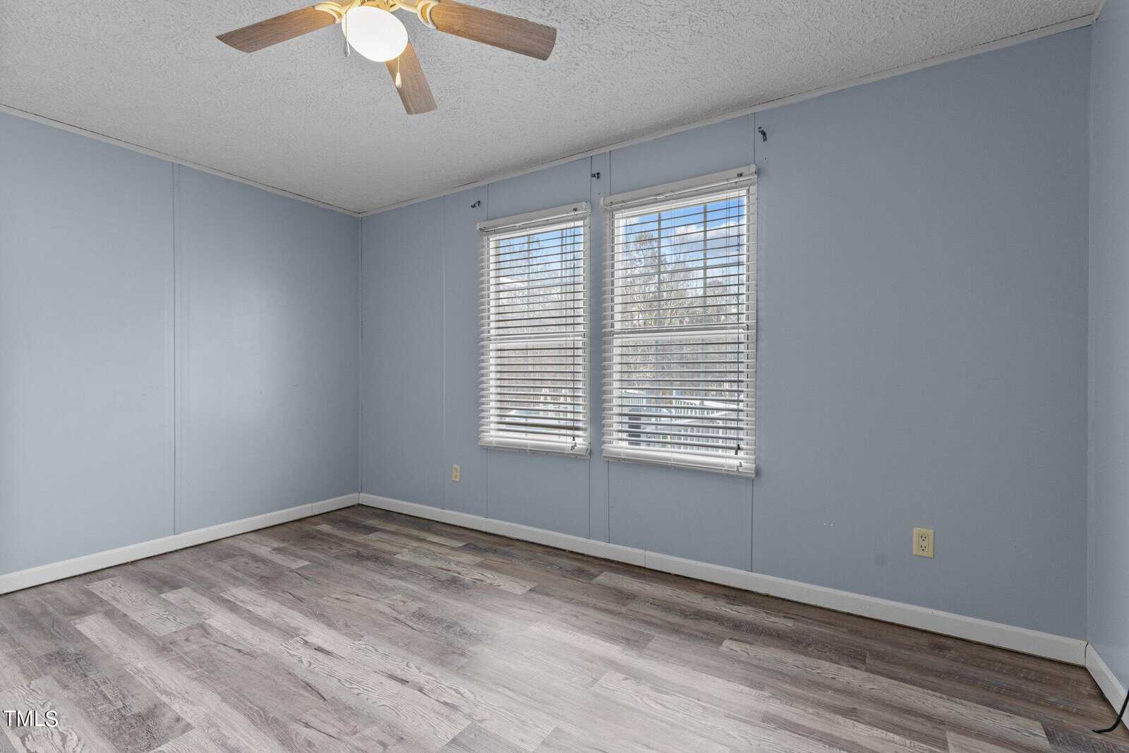 Empty room with light blue walls, wood-look flooring, two large windows with white blinds, a ceiling fan with light, and white baseboards. Natural light is coming through the windows.