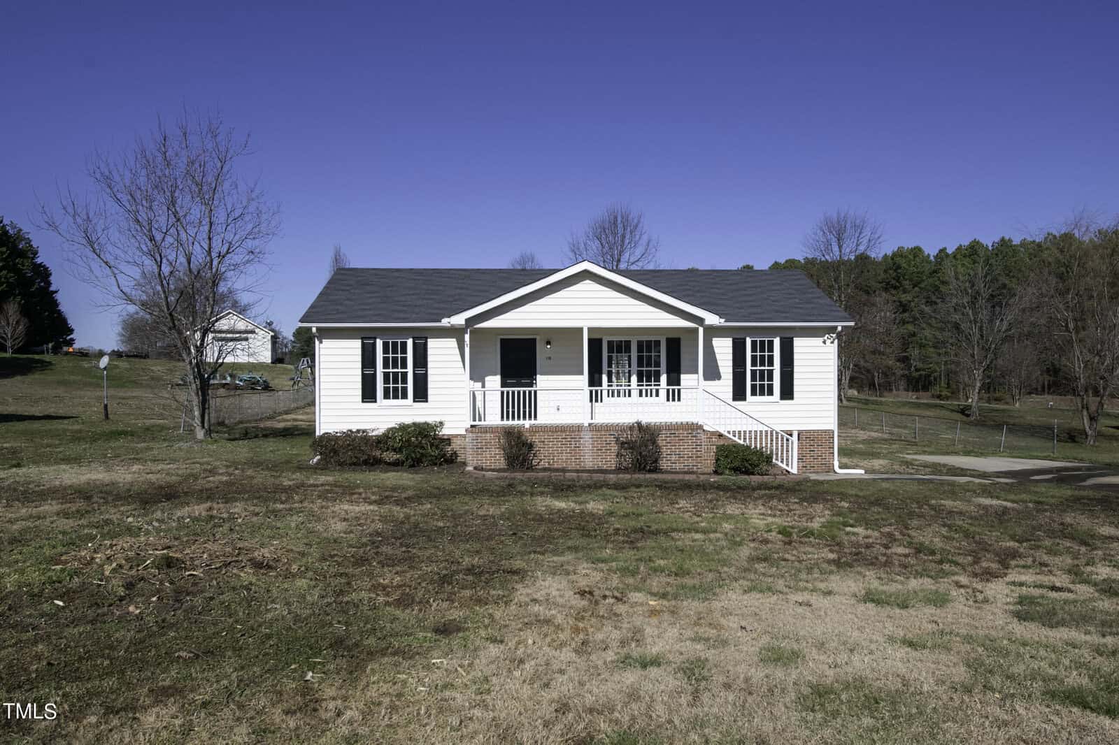A small, white, single-story house at 110 Clubview Place, complete with a charming front porch and brick steps, sits on a grassy lot in Oxford. Sparse trees are scattered around, and the sky is clear and blue in the background.
