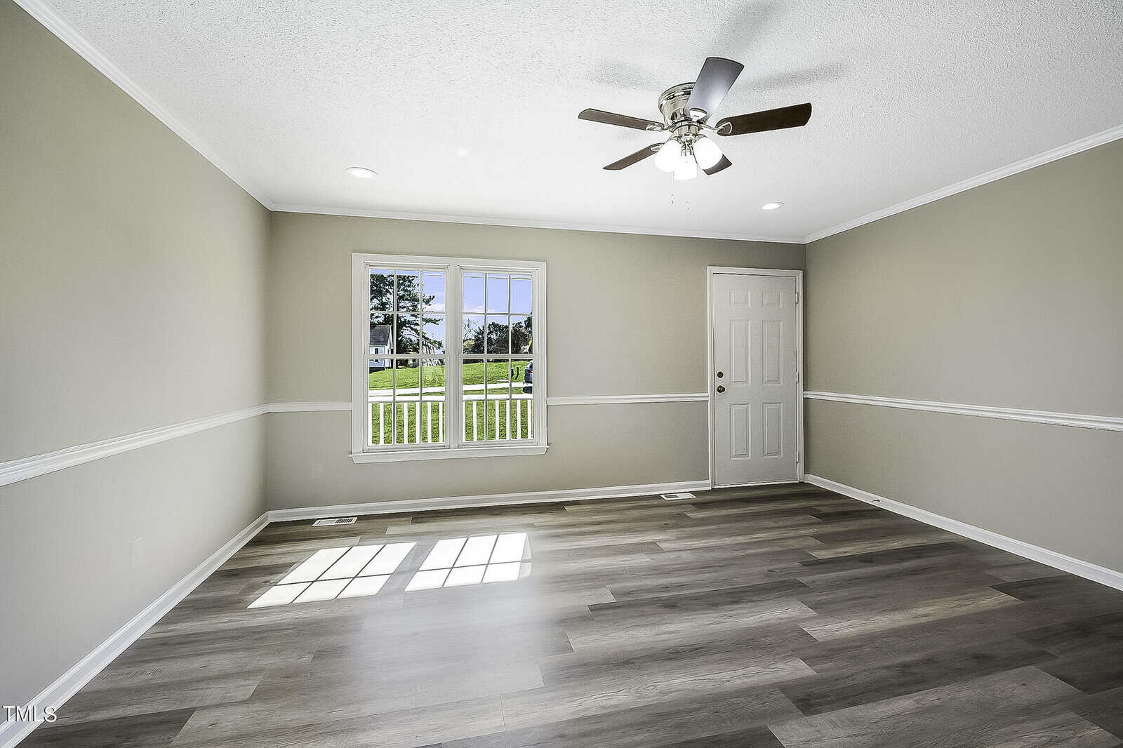 A bright, empty room with light wood flooring, neutral-colored walls, and a ceiling fan. Natural light streams through a large window and a door with a small window. The view outside shows greenery and a clear sky.
