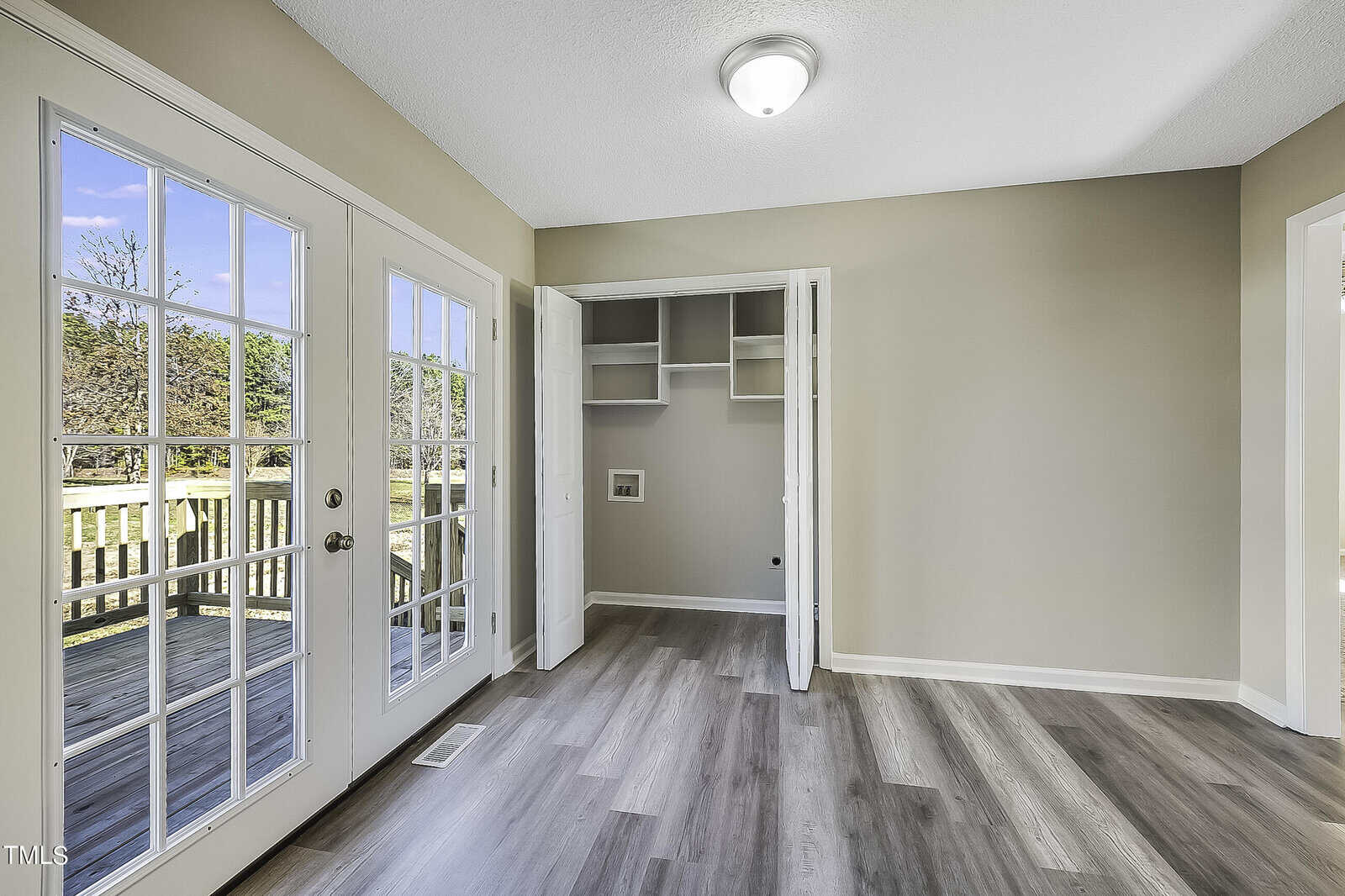 A bright room with light gray wood flooring, beige walls, and a double glass door leading to a wooden deck. An open closet with shelves is visible. Sunlight streams through the doors, illuminating the space.