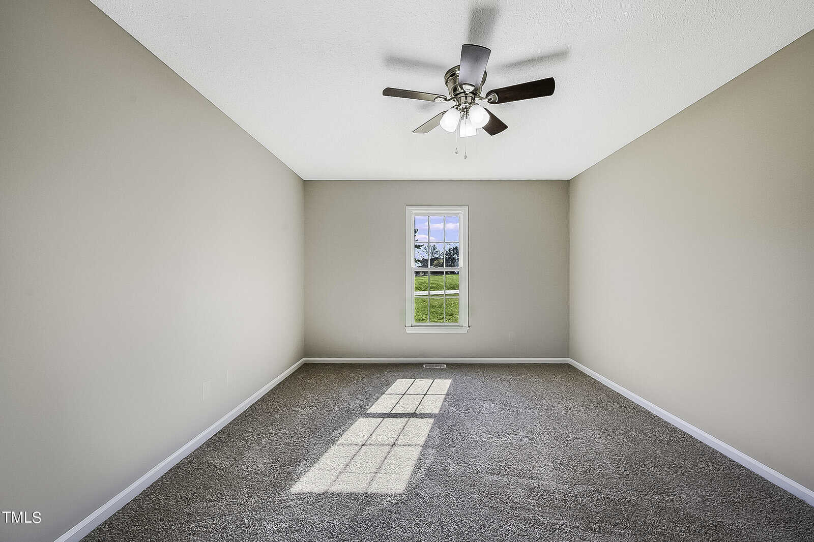 An empty room with beige walls and gray carpet, featuring a central window with a scenic view. Sunlight streams through the window, casting shadows on the floor. A ceiling fan with lights is mounted above.